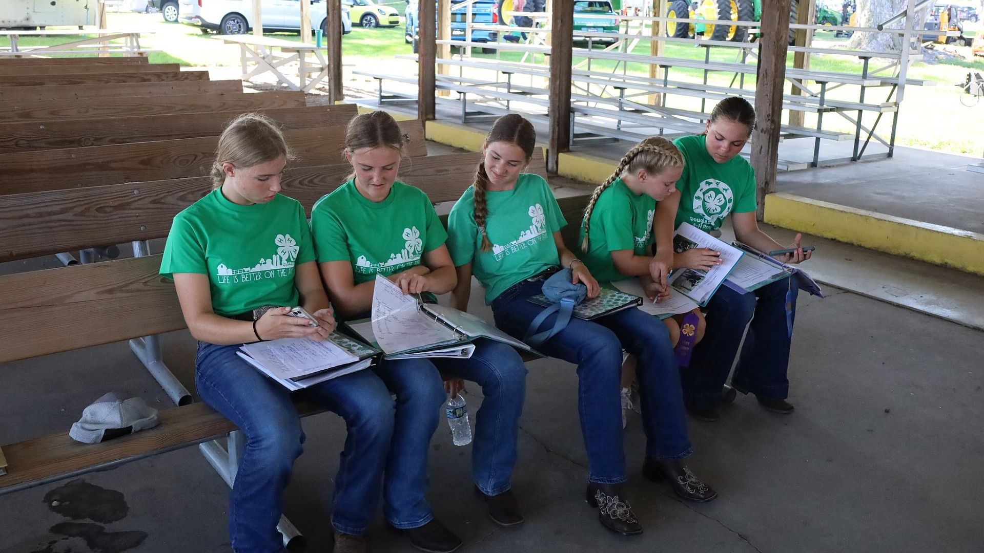 Five female 4-H exhibitors sitting on a bench in an open-air building at the Sarpy fairgrounds.