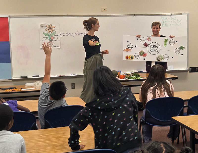 Two women teaching a class about nutrition to a group of children sitting at tables in a classroom.