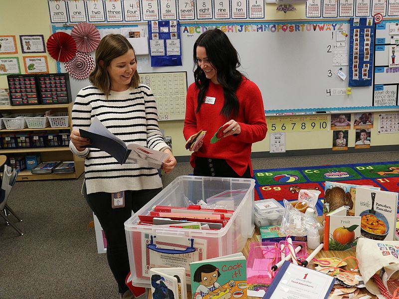 Two women looking at a notebook in a classroom with a table of books in front of them.