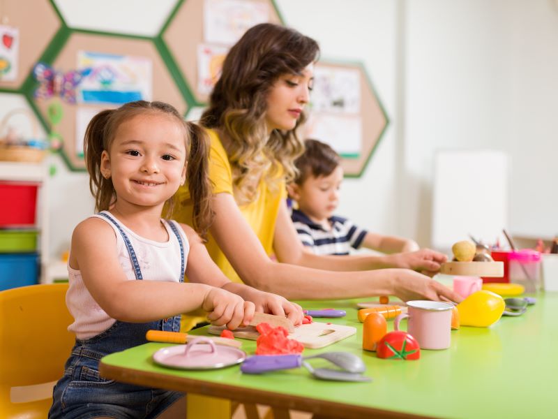 Girl role playing cutting vegetables with a woman and boy sitting at a table.