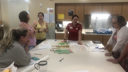 Adult women engaging around a large table in a kitchen.