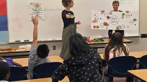 Two women teaching a class about nutrition to a group of children sitting at tables in a classroom.