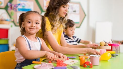 Girl role playing cutting vegetables with a woman and boy sitting at a table.