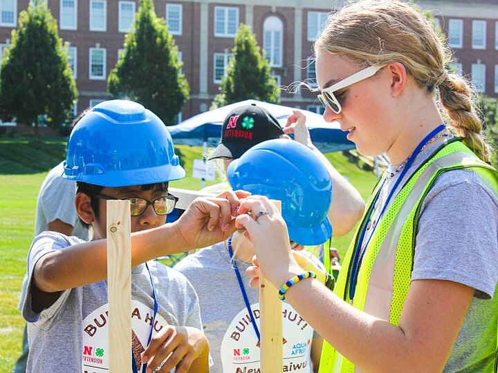 A 4-H intern assists a boy in a hard hat to construct a hut as other boys watch.