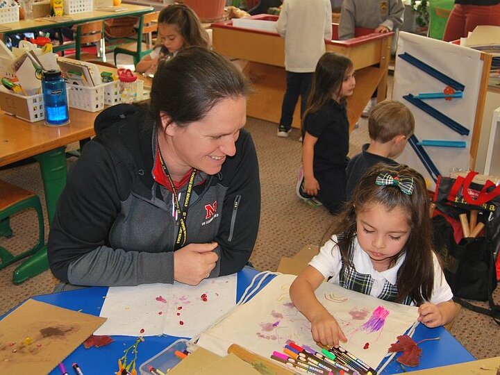 Woman is watching a girl drawing with markers at a table in a classroom.