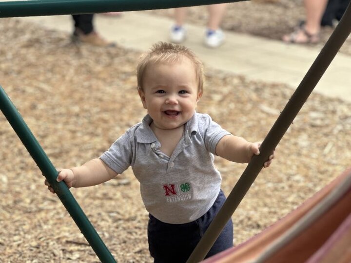 A smiling child standing on mulch outside holding on to metal pipes.