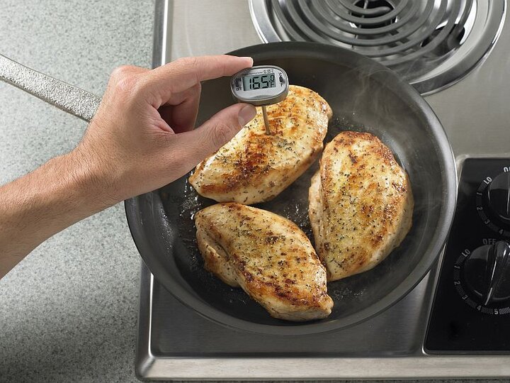 Image of a hand holding a thermometer taking the temperature of chicken cooking in a pan on a stove.