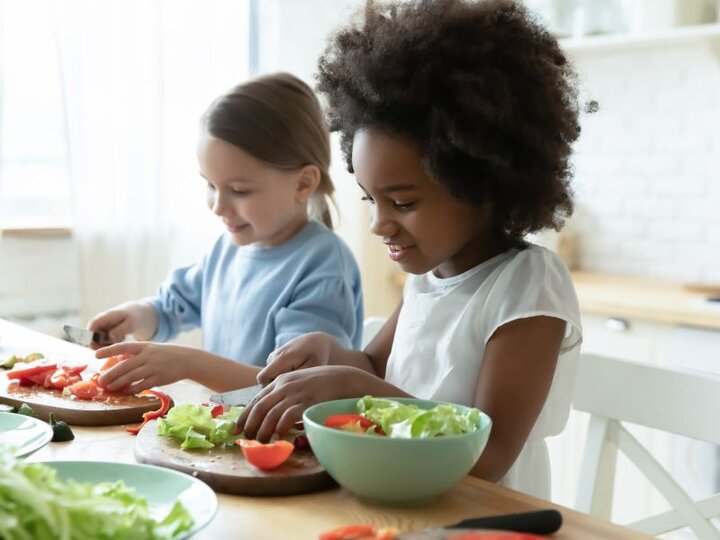 Two girls sitting at a table cutting vegetables.