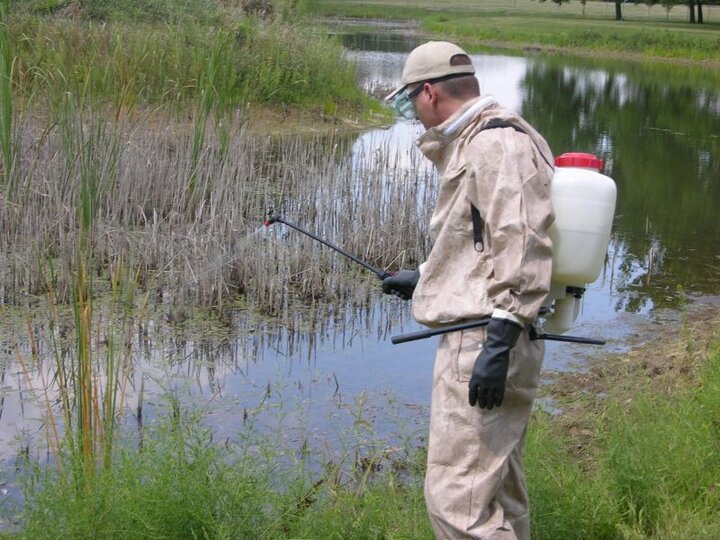 A man wearing protective clothing spraying near a body of water.